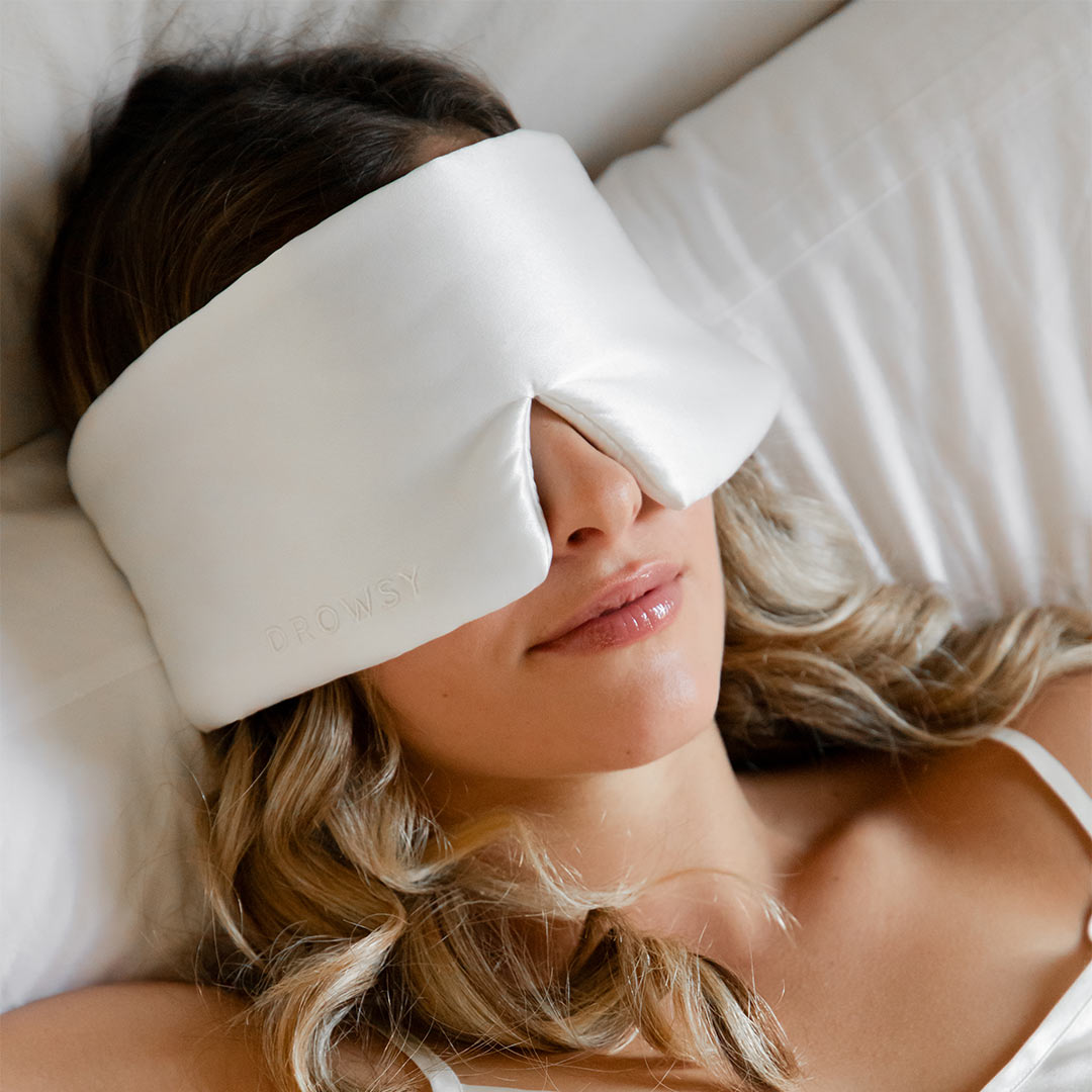 A woman with wavy blonde hair lies on a bed wearing a large, white, padded sleep mask that covers her eyes and part of her face. She appears relaxed and comfortable.