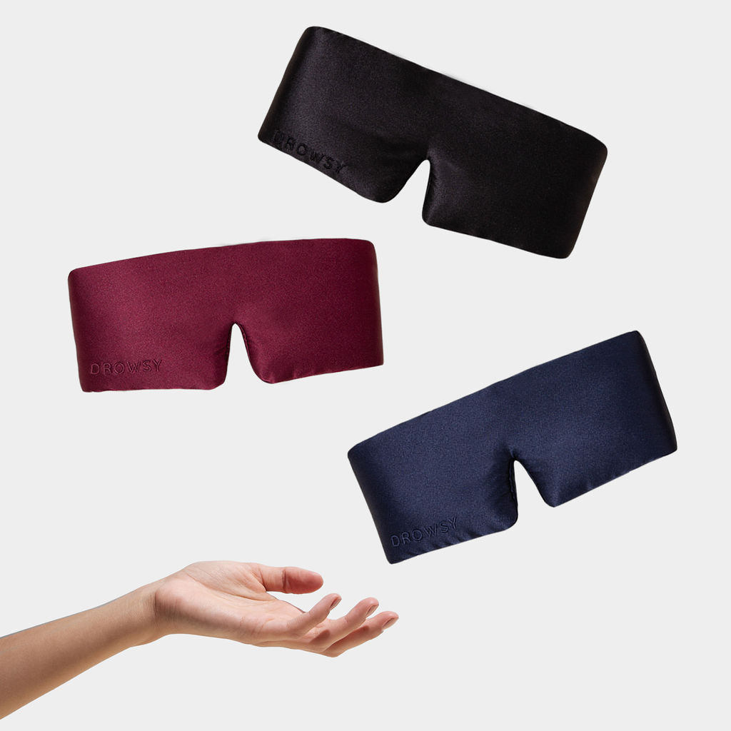 A hand reaches upward beneath three floating luxury mulberry silk masks from Drowsy’s Besties Pack—in black, burgundy, and navy—set against a plain white background.
