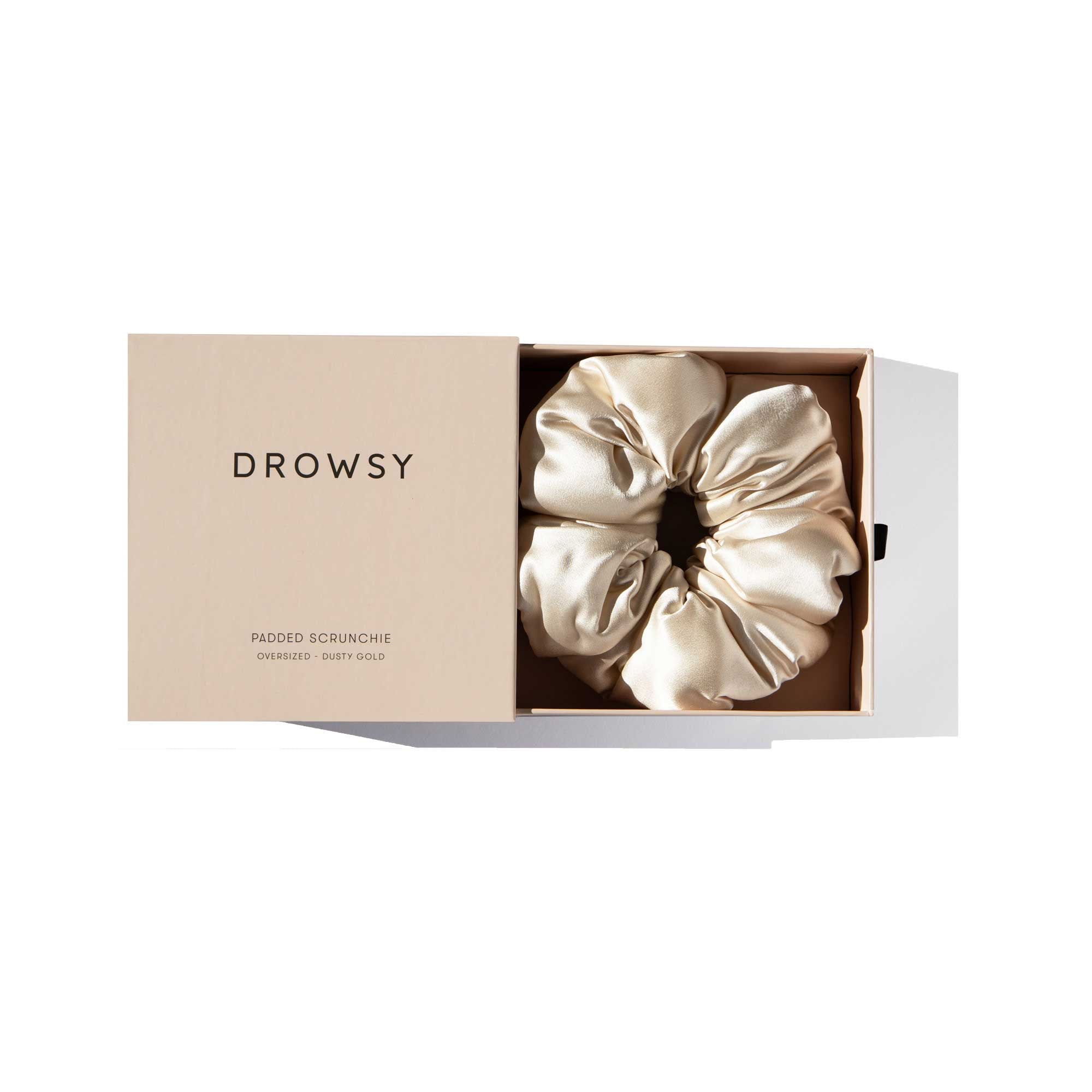 The us.drowsysleepco “Dusty Gold - Padded Scrunchie” comes in a beige box labeled “DROWSY.” Inside, the oversized, light gold silk scrunchie sits neatly on a white background—ideal for hair health.