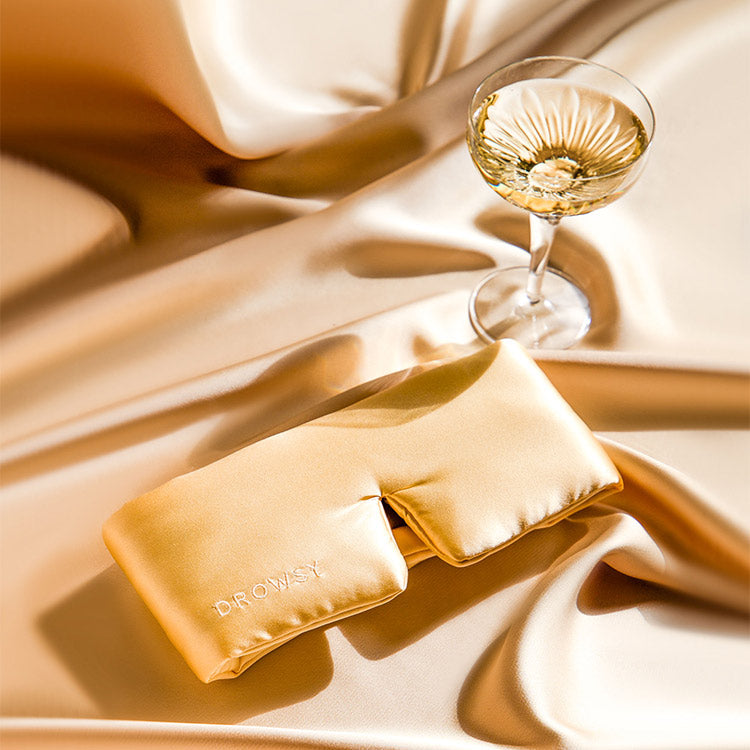A gold satin eye mask embroidered with DR. OWSY rests on matching gold satin fabric beside a glass of white wine or champagne. The scene has a luxurious and elegant feel.