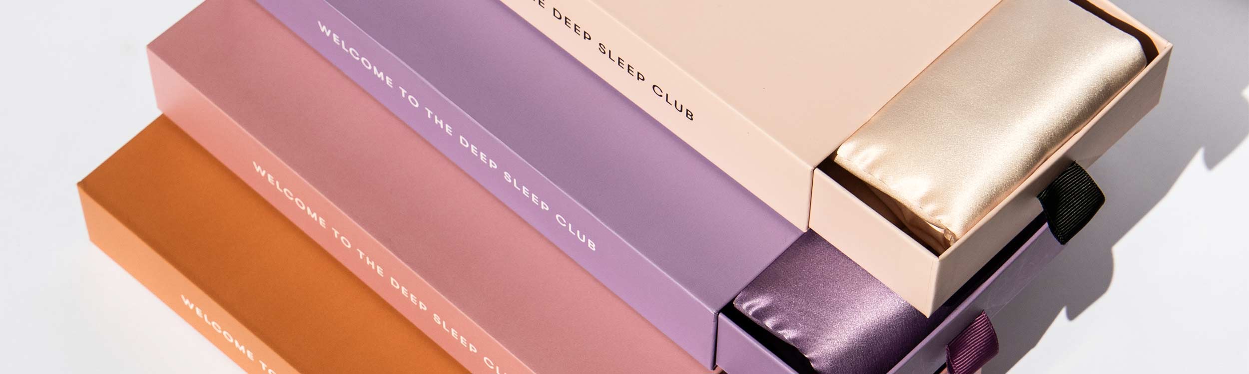 Four rectangular boxes in peach, lavender, mauve, and orange, each labeled “WELCOME TO THE DEEP SLEEP CLUB,” with drawers partially open to reveal silk sleep accessories in matching colors.