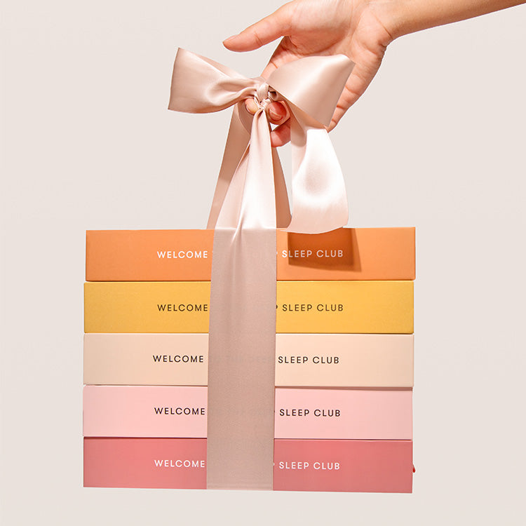 A hand holds a stack of five pastel-colored gift boxes tied together with a large, pale pink ribbon bow. The boxes are labeled “WELCOME SLEEP CLUB” and are arranged in gradient shades from orange to pink.
