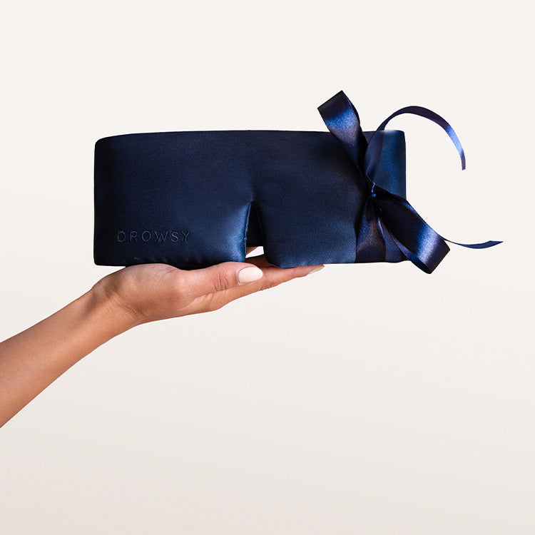 A hand with light skin and manicured nails holds a dark blue silk sleep mask with a large matching ribbon and the word DROWSY embossed on it, against a plain light background.