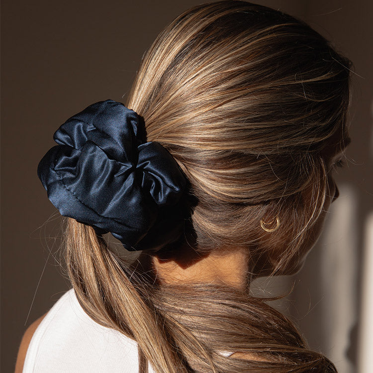 A woman with light brown hair wears a large, dark blue satin scrunchie in a low ponytail. She is wearing a white top and a small gold hoop earring, facing away from the camera in soft sunlight.