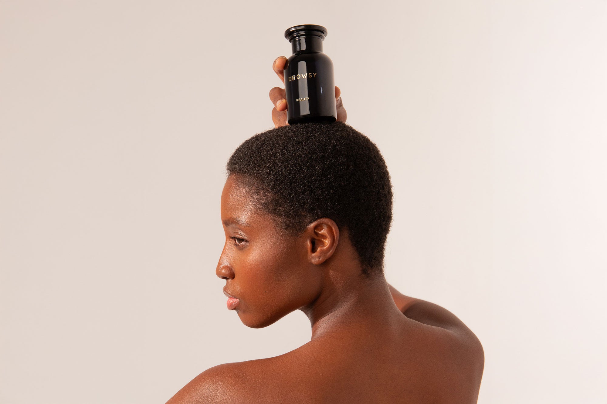 A person with short hair holds a black bottle on their head, posing against a neutral background with their shoulder and profile visible.