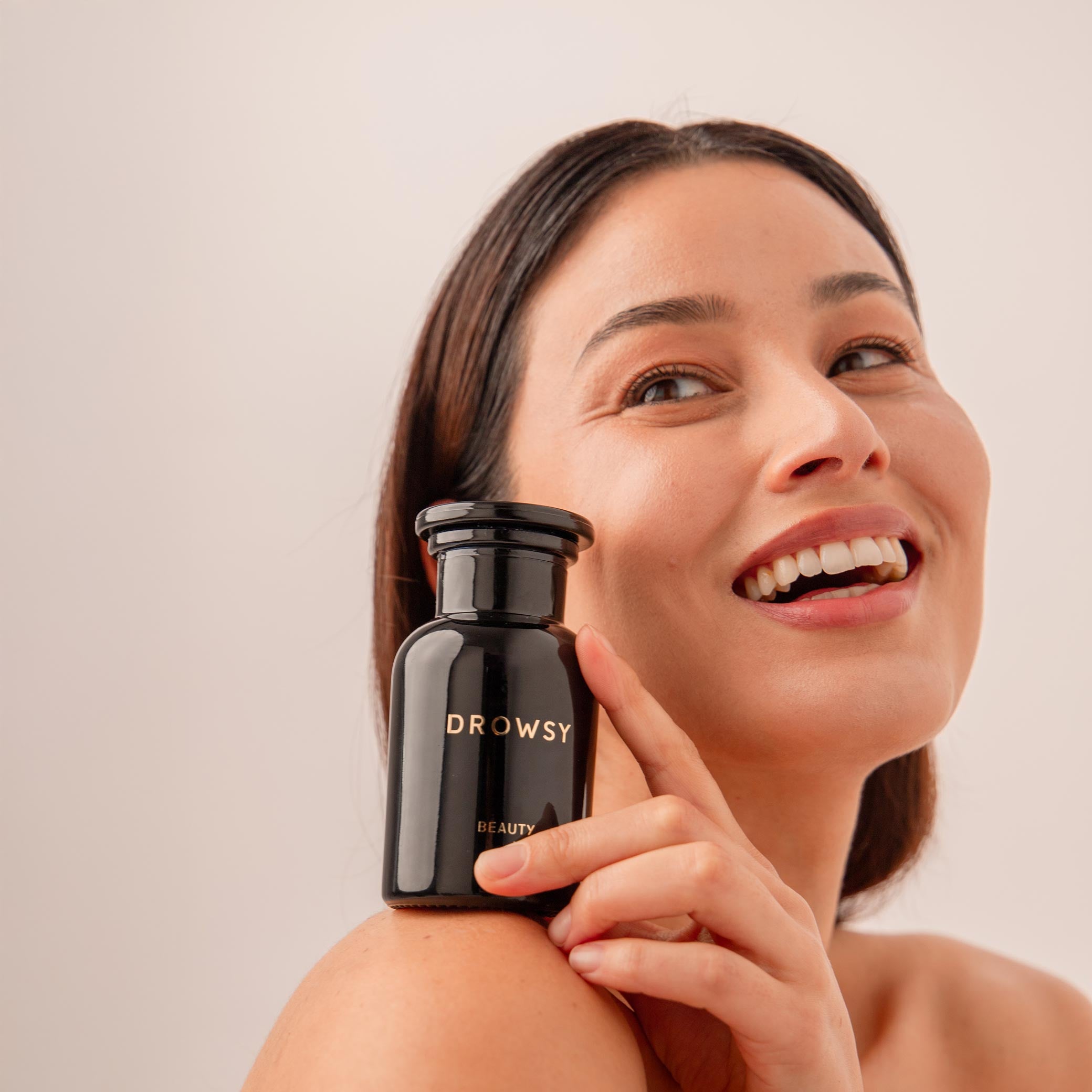 A smiling woman holds a black us.drowsysleepco Supplements - Beauty bottle close to her face, her skin glowing with a youthful complexion against a simple light background.