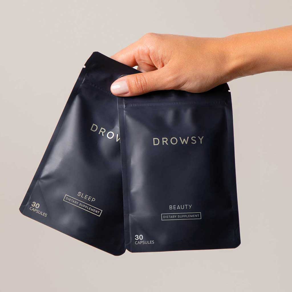 A hand holds two black packets labeled Drowsy. One packet says Sleep and the other Beauty. Each contains 30 dietary supplement capsules.