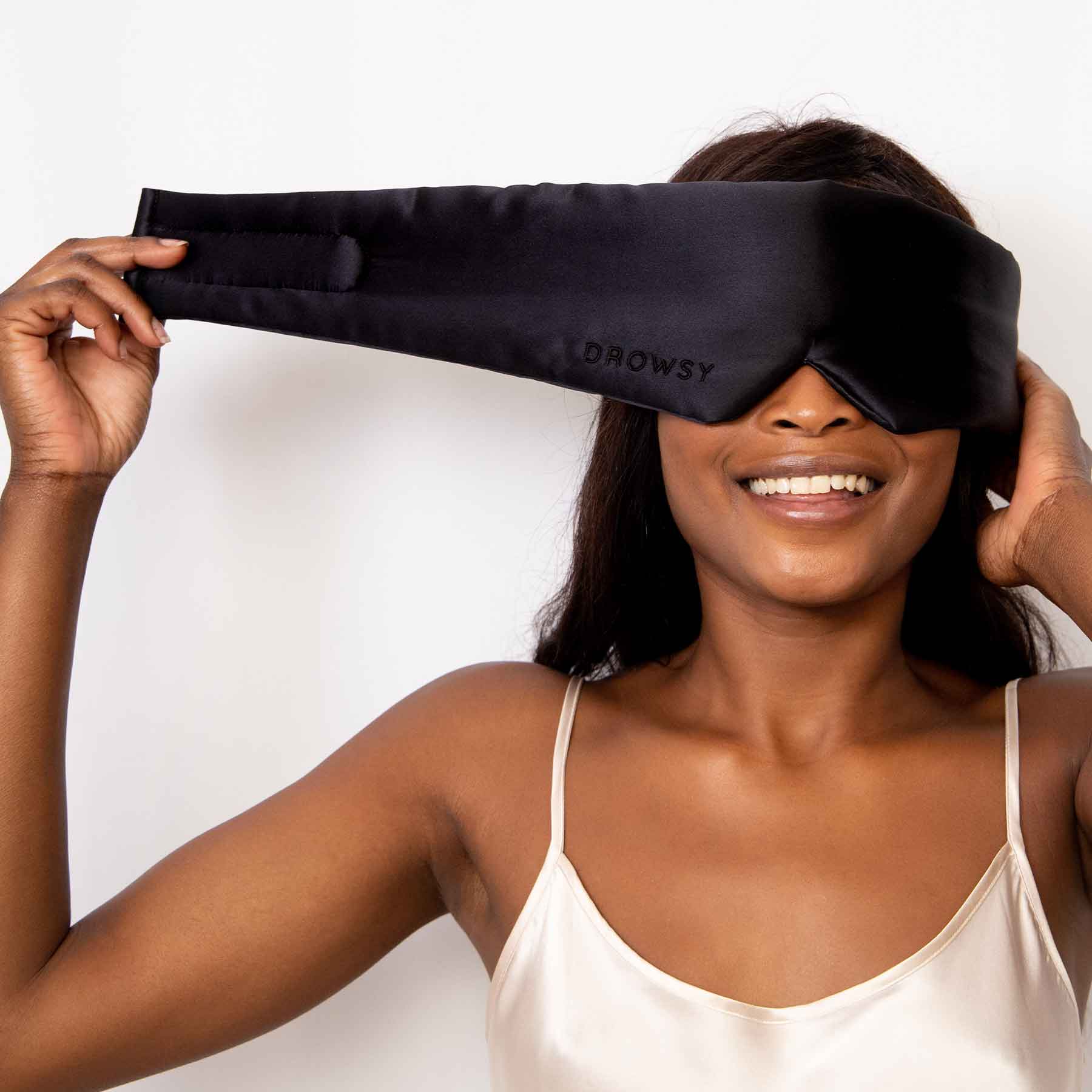 A smiling woman wearing a cream-colored camisole adjusts a large black sleep mask labeled DROWSY over her eyes against a plain white background.