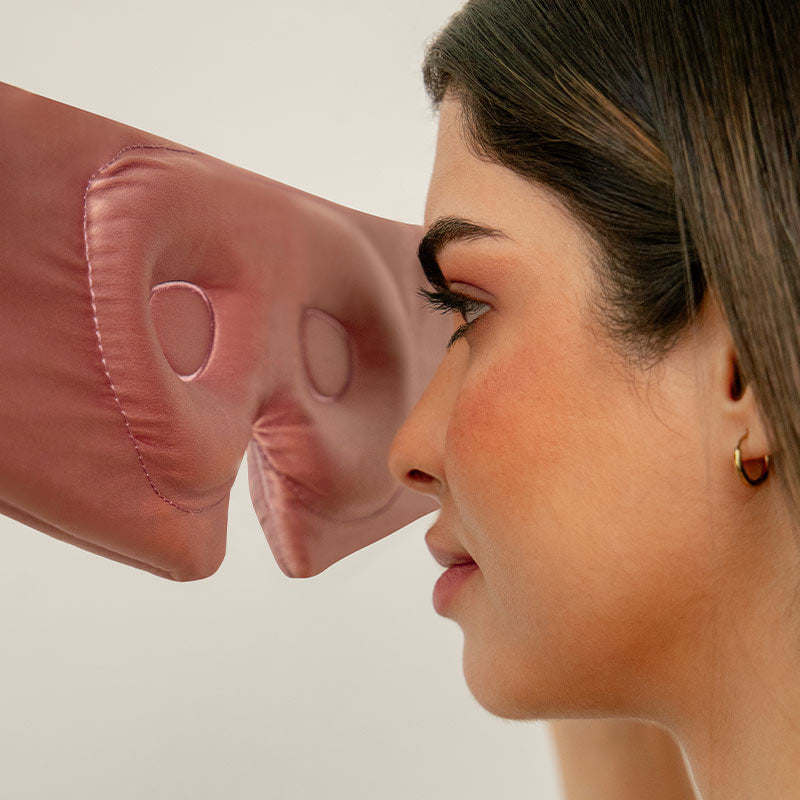 A woman with long brown hair faces a pink, padded eye mask with large eye holes, holding it close to her face as if about to put it on. The background is simple and neutral.