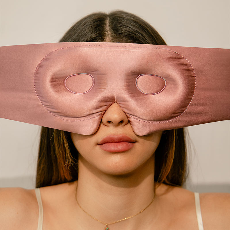 A woman with long brown hair wears a pink satin sleep mask incorrectly over her eyes, with the mask’s eye holes above her eyes and her facial expression neutral.