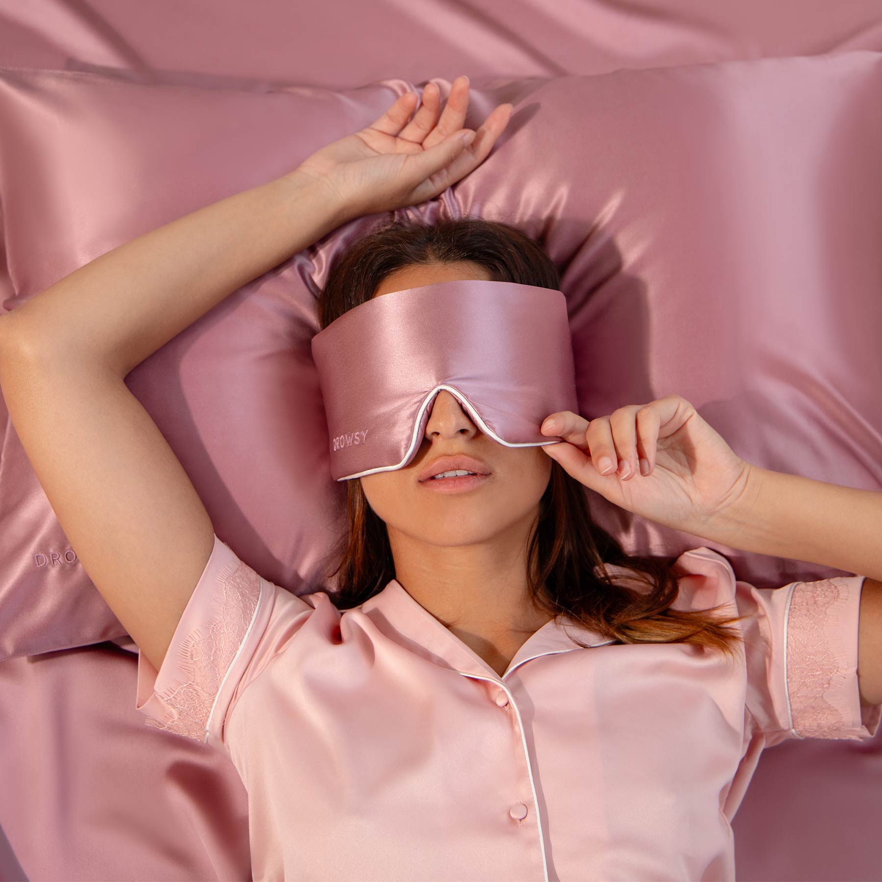 A woman in pink satin pajamas relaxes on a matching bed, adjusting her Damask Rose - Pipe Dreams Edition silk sleep mask by us.drowsysleepco, enjoying total darkness and comfort from its blackout design and adjustable strap.