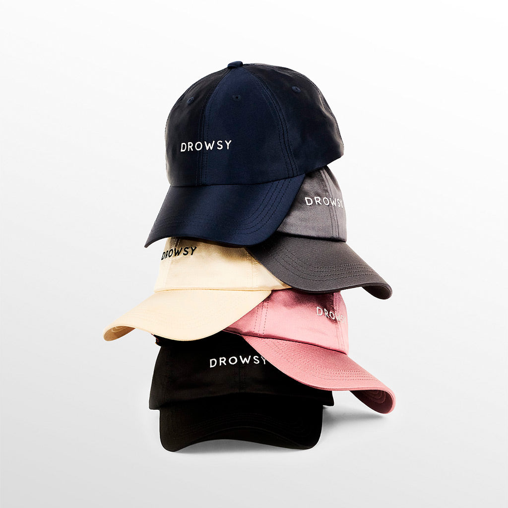 A stack of five baseball caps in navy blue, gray, beige, pink, and black, each embroidered with the word DROWSY on the front, against a plain white background.