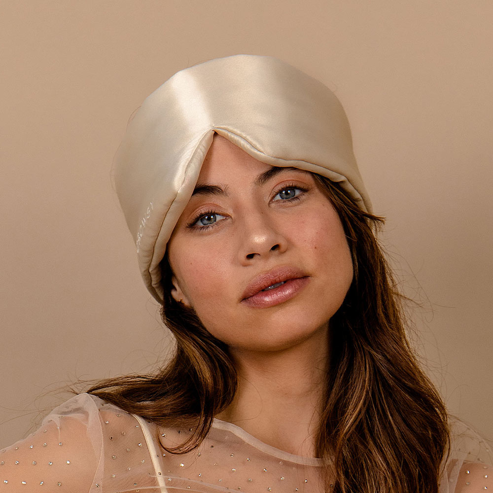 A woman with long brown hair and light eyes wears the Sensual Sleep Bundle - Dusty Gold satin turban by us.drowsysleepco, styled with a sheer beige top against a plain beige background.
