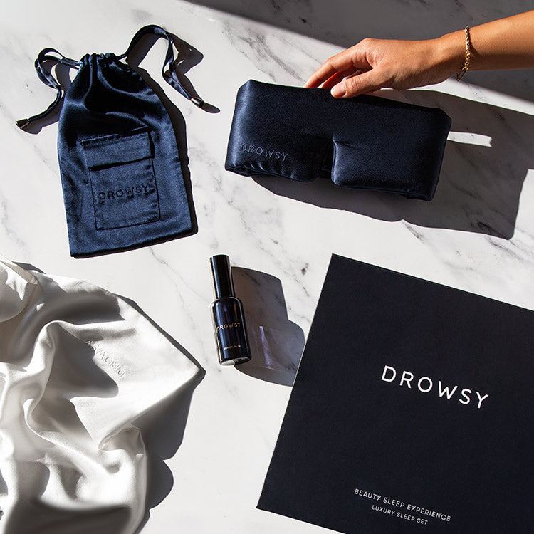 A hand holds a black silk sleep mask next to a matching drawstring pouch, a spray bottle, a white silk pillowcase, and a black Drowsy gift box on a marble surface.
