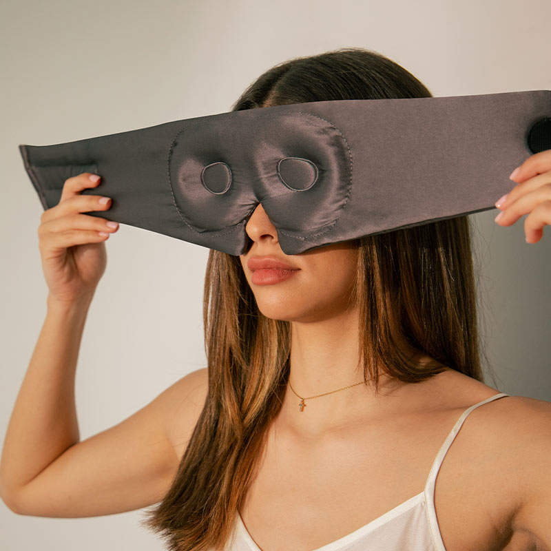 A woman with long brown hair wears a white camisole and holds a black, padded sleep mask with eye cutouts over her eyes.