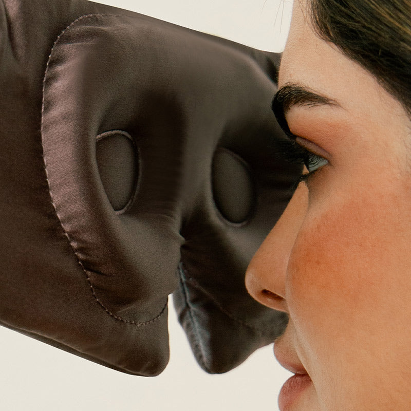 Close-up of a woman’s face wearing a brown silky sleep mask with deep eye cups, designed to avoid contact with her eyes and eyelashes. The background is plain and light-colored.