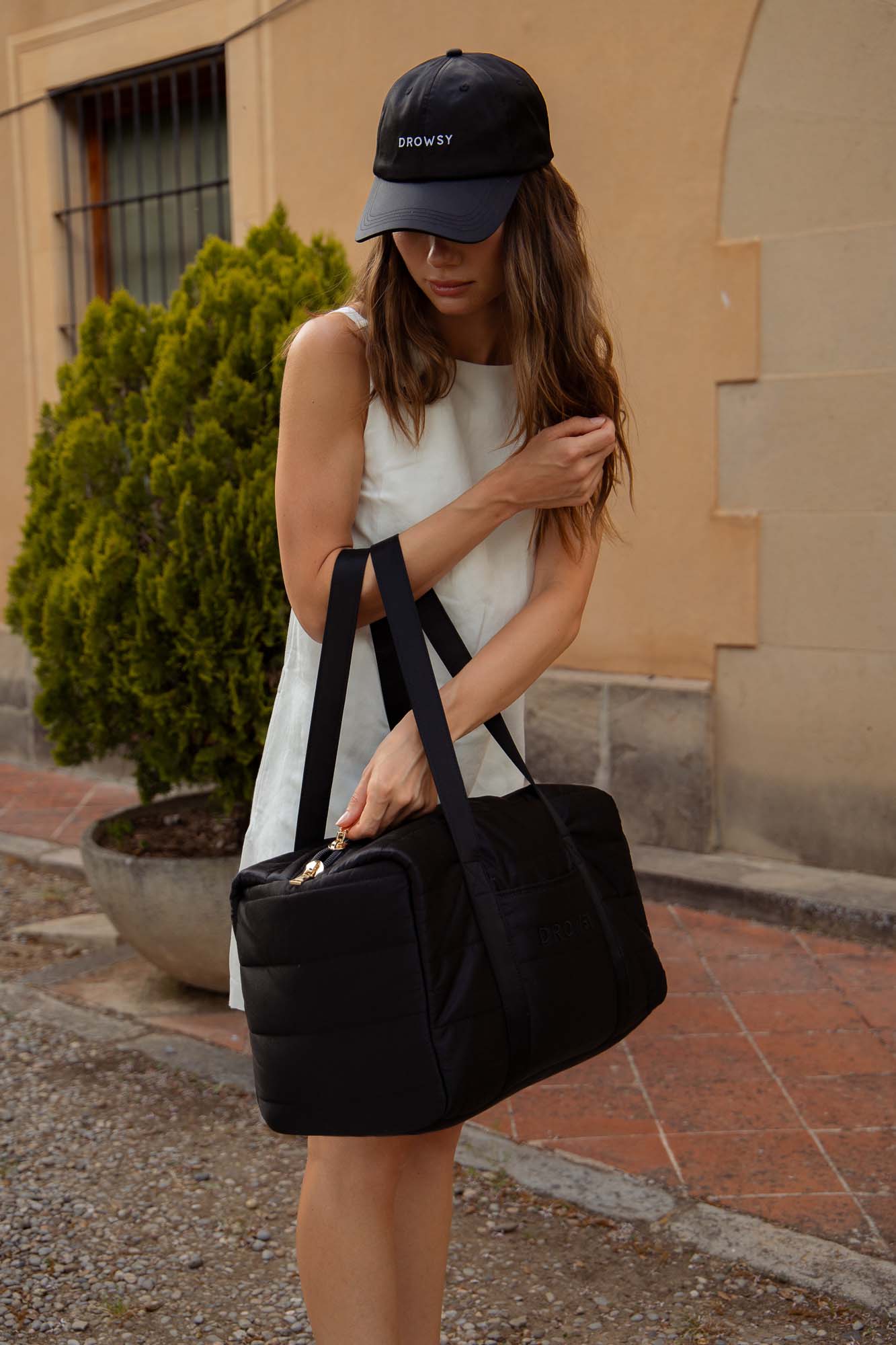 A woman in a sleeveless white dress and black cap adjusts her hair while carrying a large black duffel bag outdoors, with a beige wall and a potted shrub in the background.