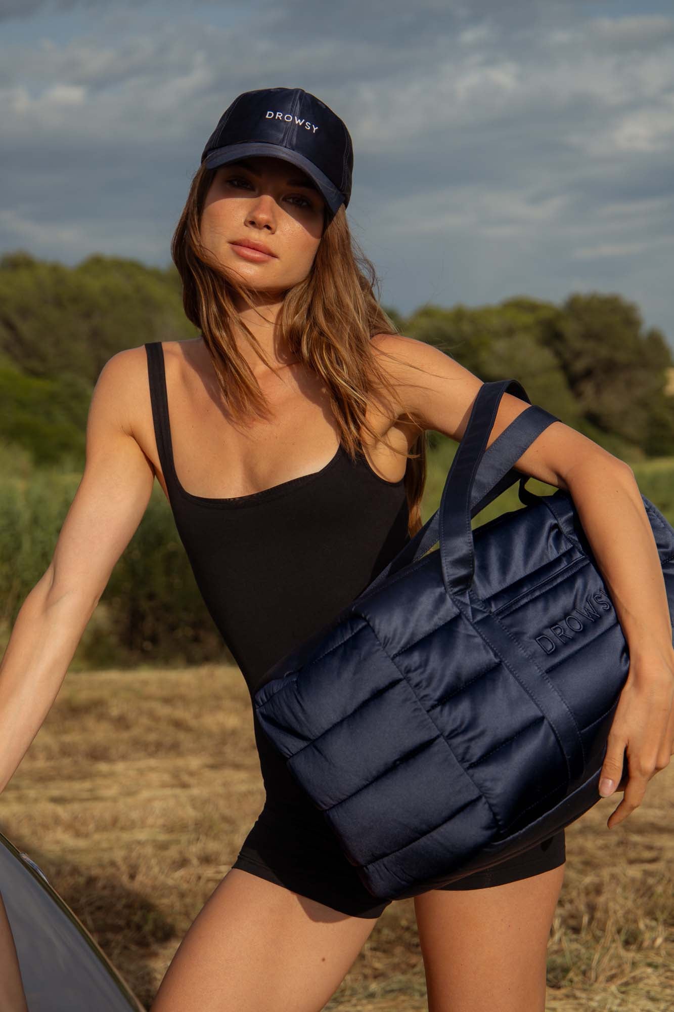 A woman wearing a black sleeveless romper and a navy Drowsy cap stands outdoors, holding a matching navy quilted bag. She poses confidently, with greenery and a cloudy sky in the background.