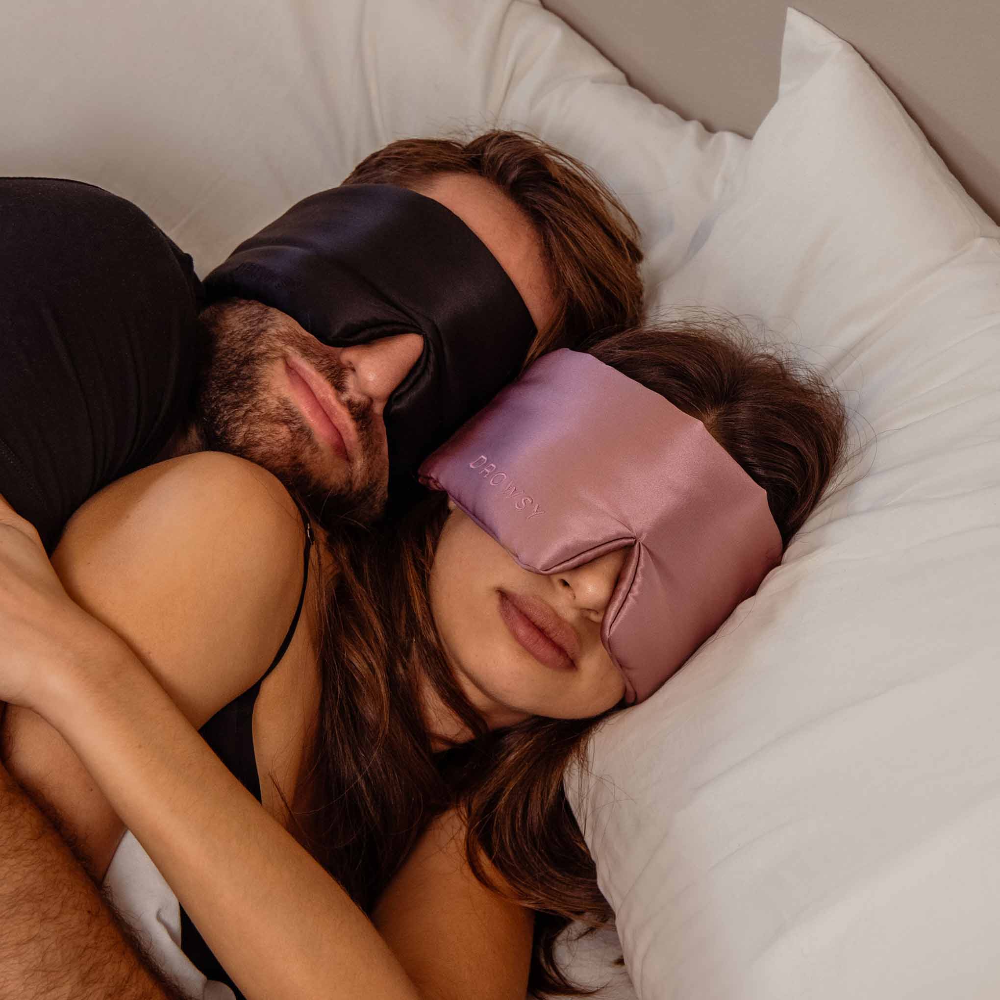 A couple cuddles in bed on white pillows, each wearing a Drowsy Lovers Pack luxury silk mask—one black, one pink—enjoying restful beauty sleep.