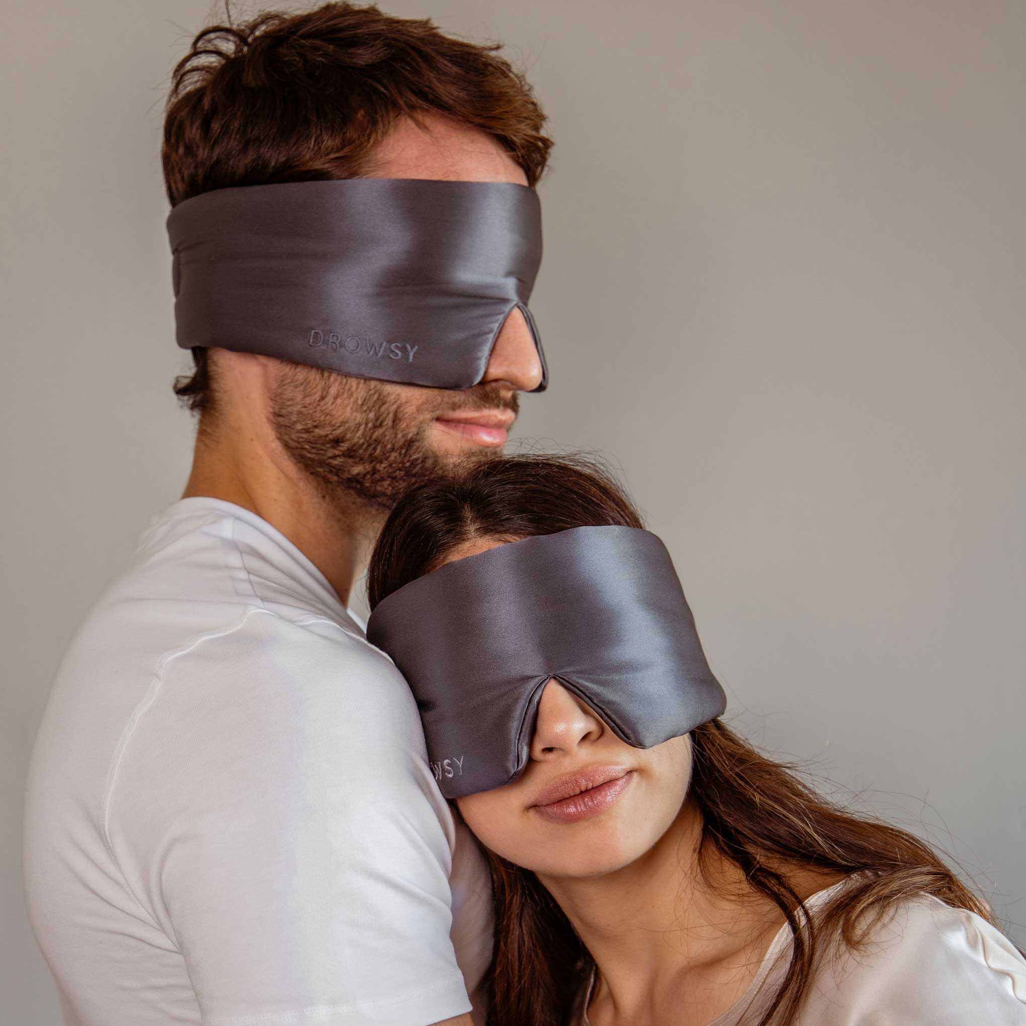 A man and woman in white shirts relax together against a plain background, both wearing Drowsy’s Lovers Pack: large, dark gray luxury silk masks. The woman leans on the mans chest, savoring true beauty sleep.