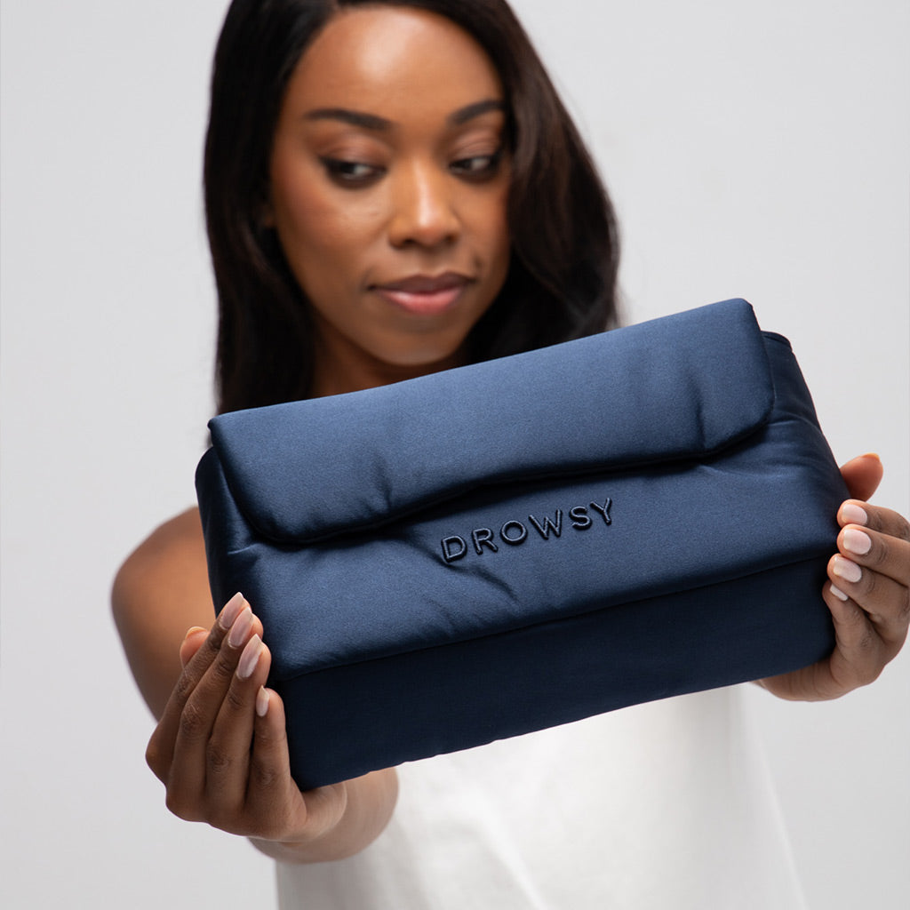 A woman in a white top holds out the Drowsy Silk Necessaire - Midnight Blue by Drowsy, a padded silk pouch with DROWSY embroidered on it, smiling slightly against a plain background.