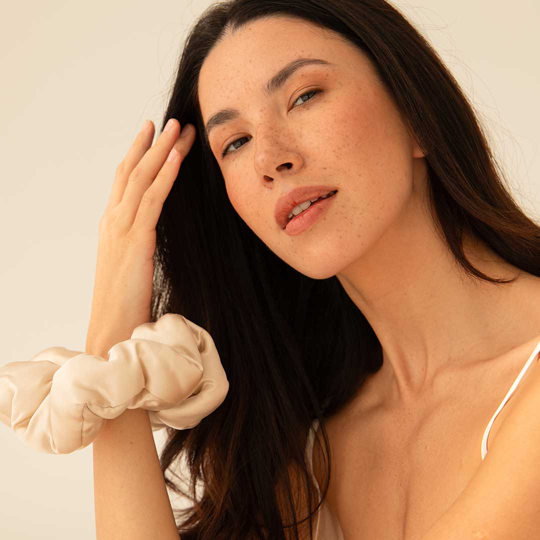 A woman with long brown hair and natural makeup touches her hair with one hand. She wears a light-colored scrunchie on her wrist and a white spaghetti strap top. The background is neutral and softly lit.
