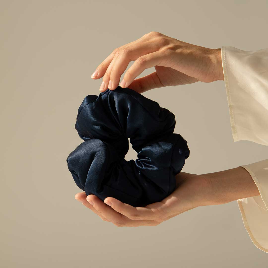 Two hands gently hold a large, dark blue satin scrunchie against a neutral background. The person is wearing a light-colored, long-sleeved garment.