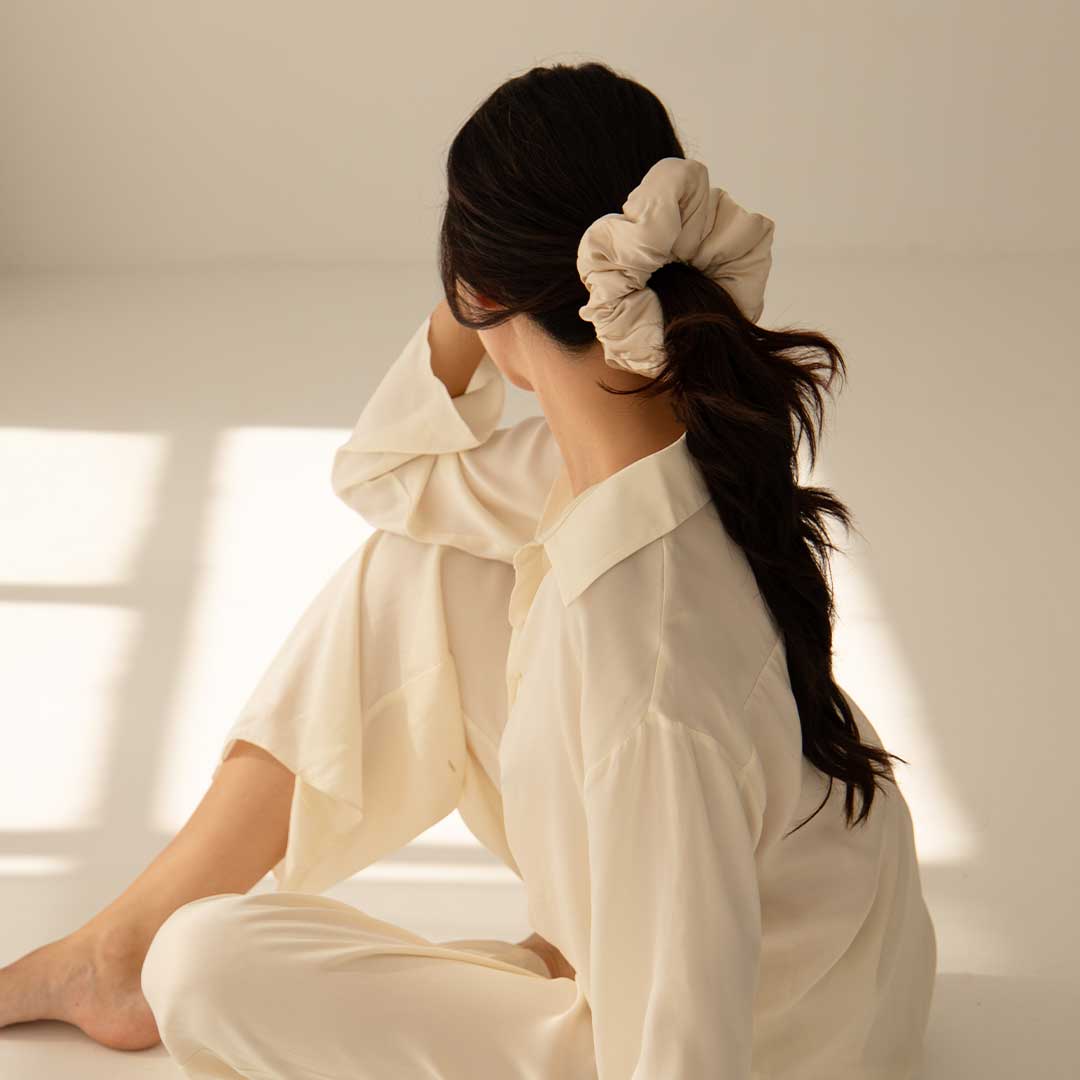 A woman with long dark hair tied in a beige scrunchie sits on the floor in soft natural light, wearing cream-colored pajamas and looking away from the camera.