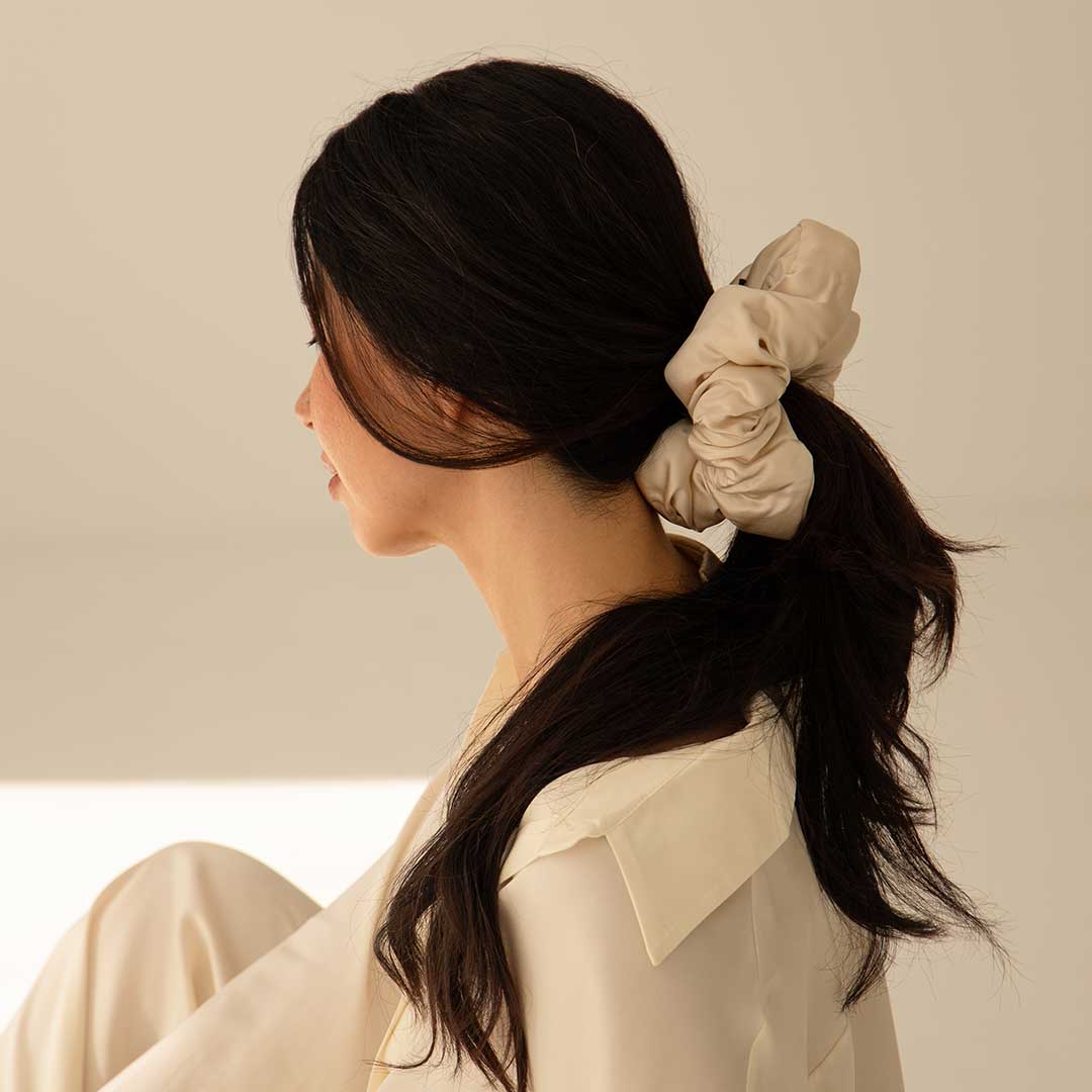 A woman with long dark hair in a low ponytail secured by the us.drowsysleepco Dusty Gold - Padded Scrunchie wears a cream blouse, sitting sideways in soft light and capturing an effortless, healthy hair style.