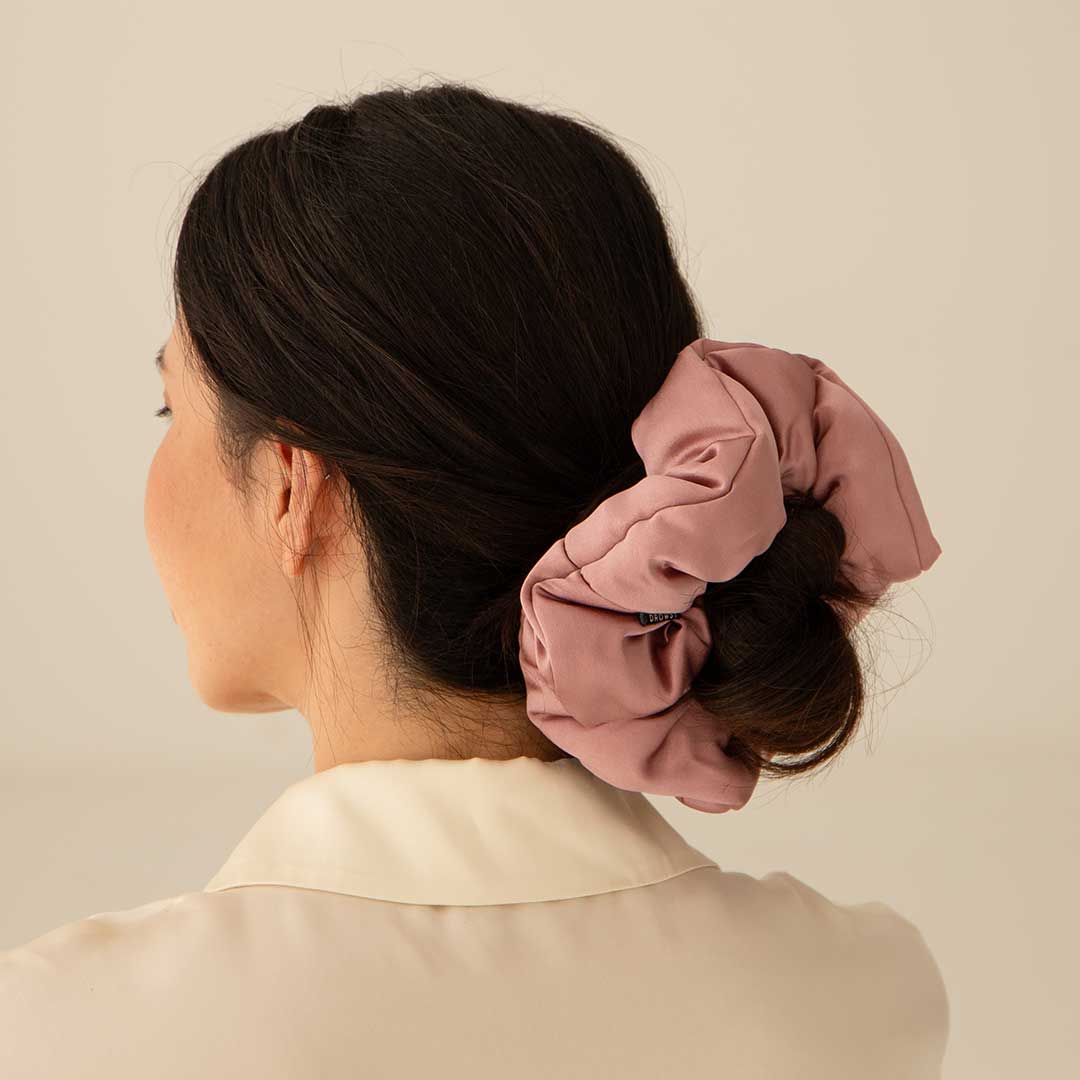 A woman with dark hair in a low bun, secured by the us.drowsysleepco Damask Rose - Padded Scrunchie, is shown from behind and slightly to the side, wearing a cream collared shirt against a plain background.