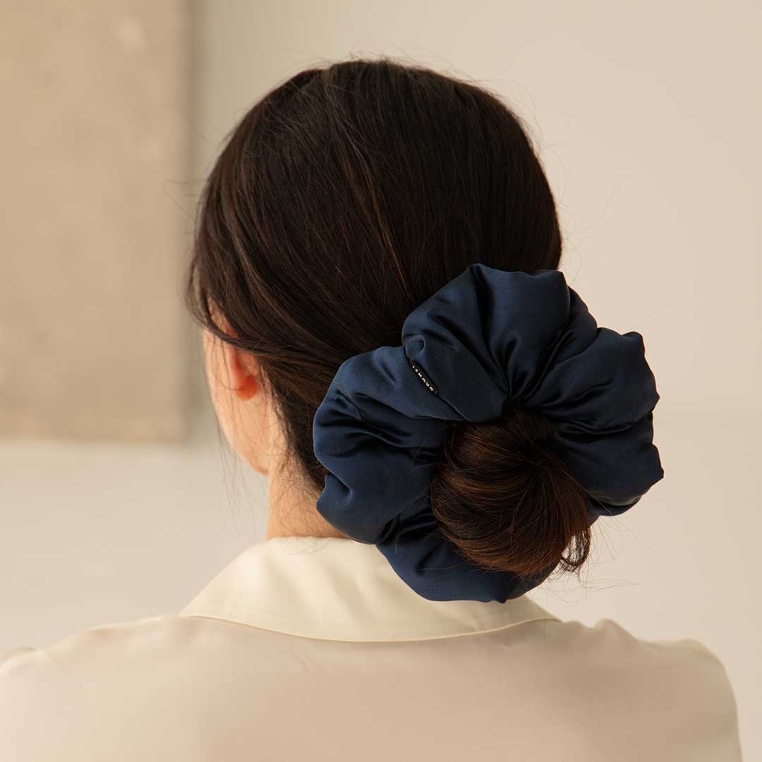 A woman with dark hair styled in a low bun, secured with a large navy blue scrunchie, is seen from behind. She is wearing a cream-colored blouse.