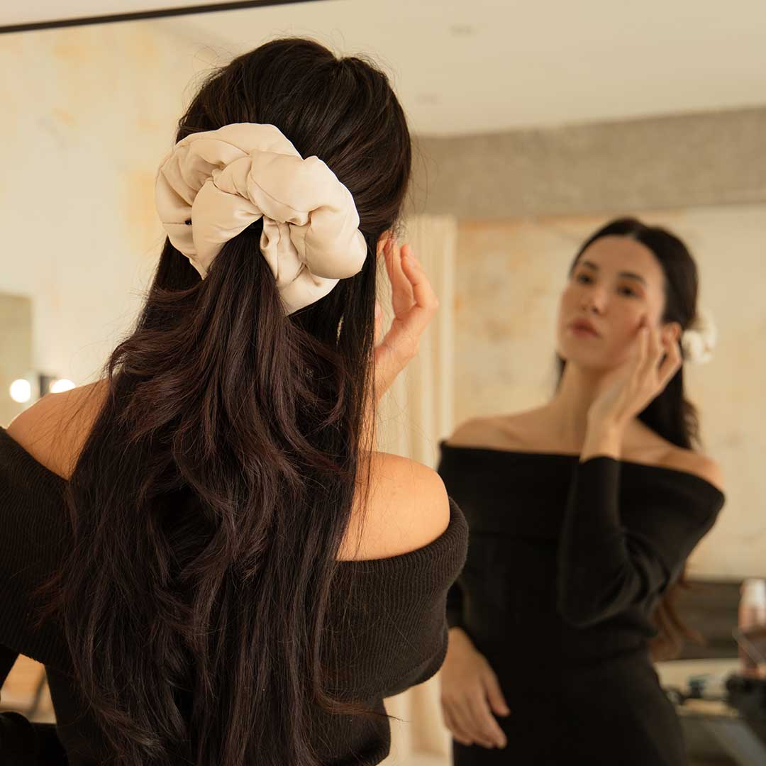 A woman with long dark hair, wearing an off-the-shoulder black top and a large light scrunchie, stands in front of a mirror touching her face and looking at her reflection.