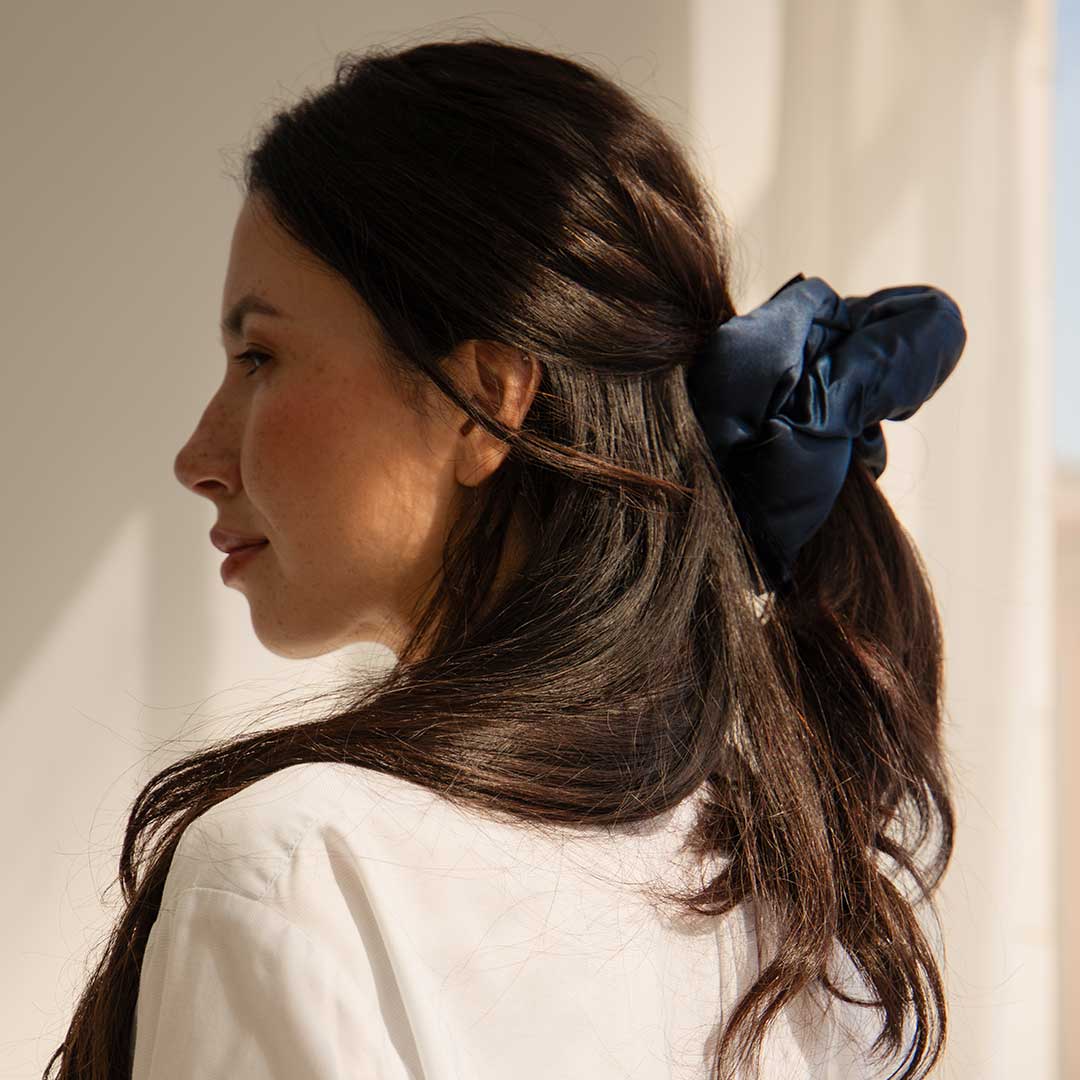Woman with long dark hair wearing a white shirt and a navy blue scrunchie, looking to the left in soft natural light.