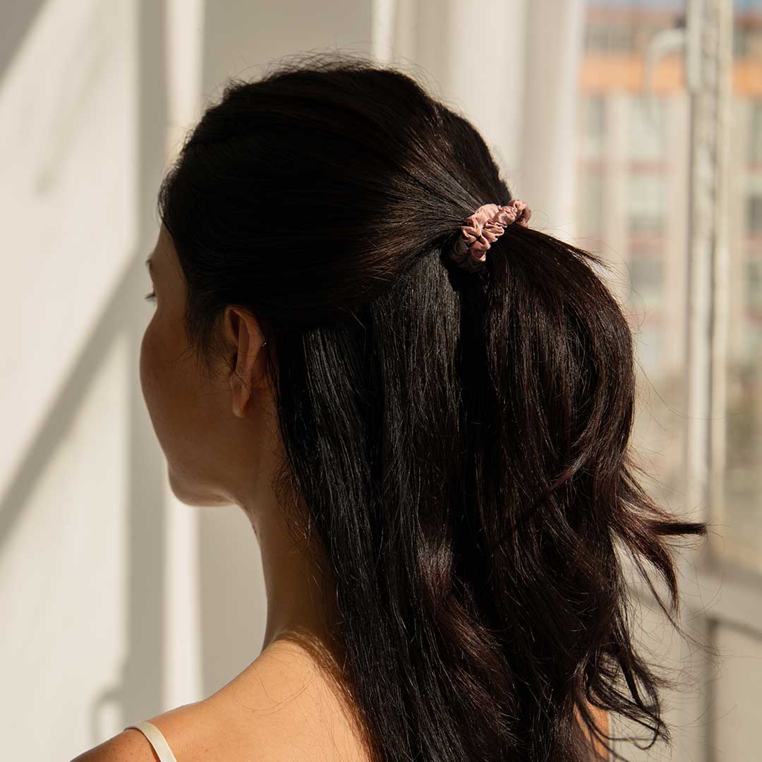 A woman with long dark hair, tied back with a pink scrunchie, stands in sunlight near a window, facing away from the camera.
