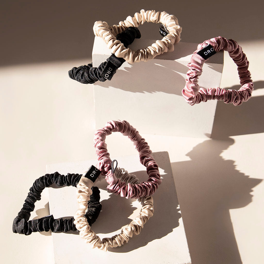 The us.drowsysleepco Skinny Scrunchie Set—featuring Rose, Dusty, and Black shades—is crafted from luxurious 22 momme silk. Arranged on white blocks, each scrunchie has a black tag and smooth texture for optimal hair health.