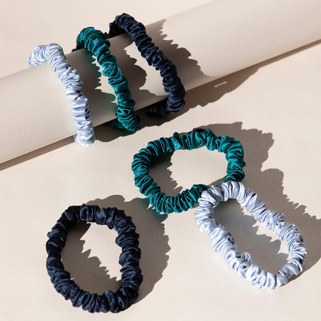 The us.drowsysleepco Skinny Scrunchie Set - Blue & Green Shades features six skinny 22 momme silk scrunchies in navy, teal, and light blue, arranged on a beige surface with three on a rolled sheet and three lying flat.