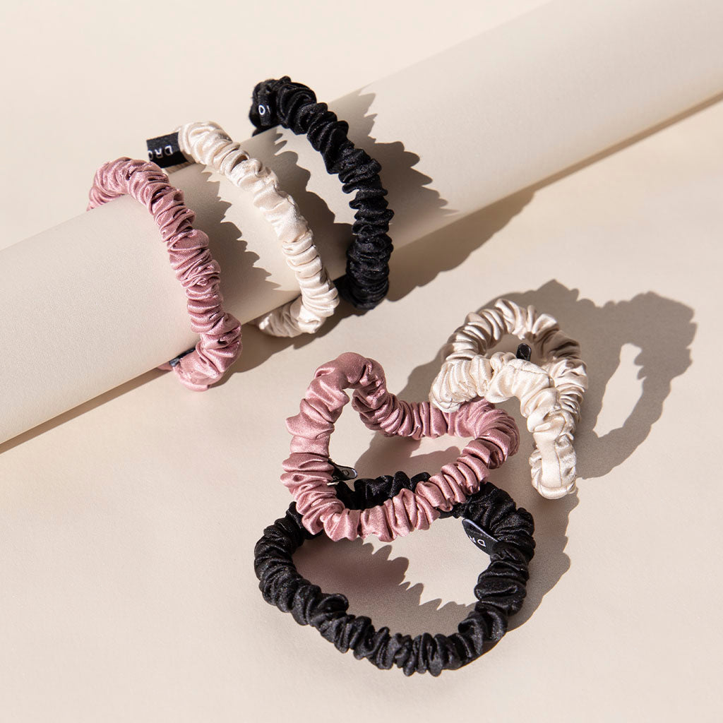 The us.drowsysleepco Skinny Scrunchie Set in Rose, Dusty, and Black includes six 22-momme silk scrunchies shown on a white cylinder and beige surface—ideal for hair health.