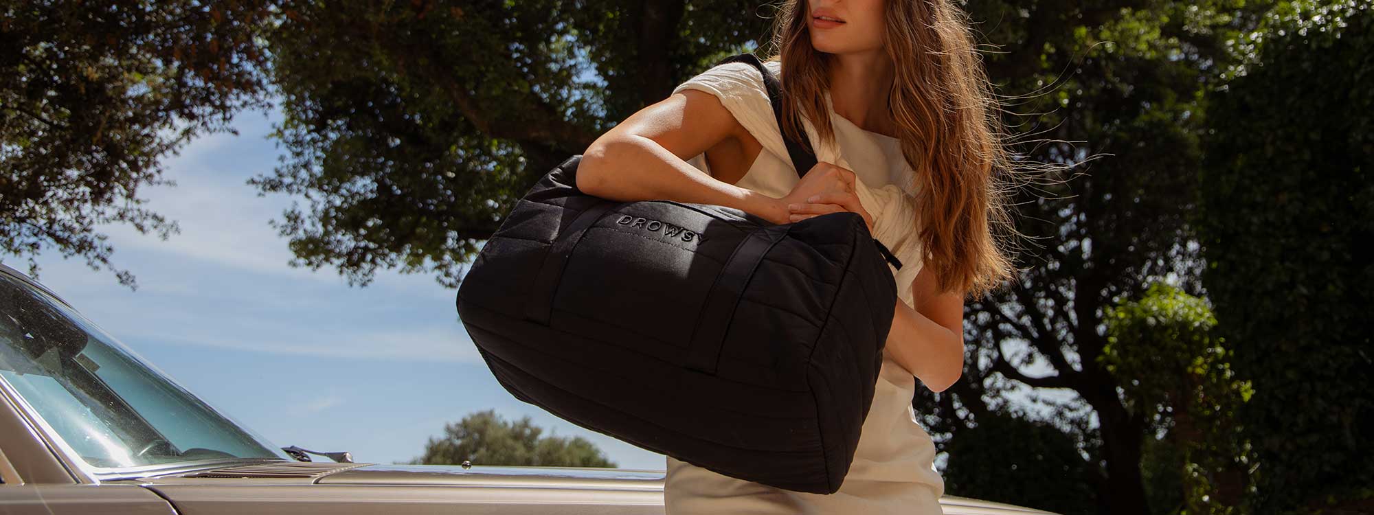 A woman in a light dress stands outdoors, holding a large black duffel bag with CROWNE written on it, near a classic white car and surrounded by greenery.