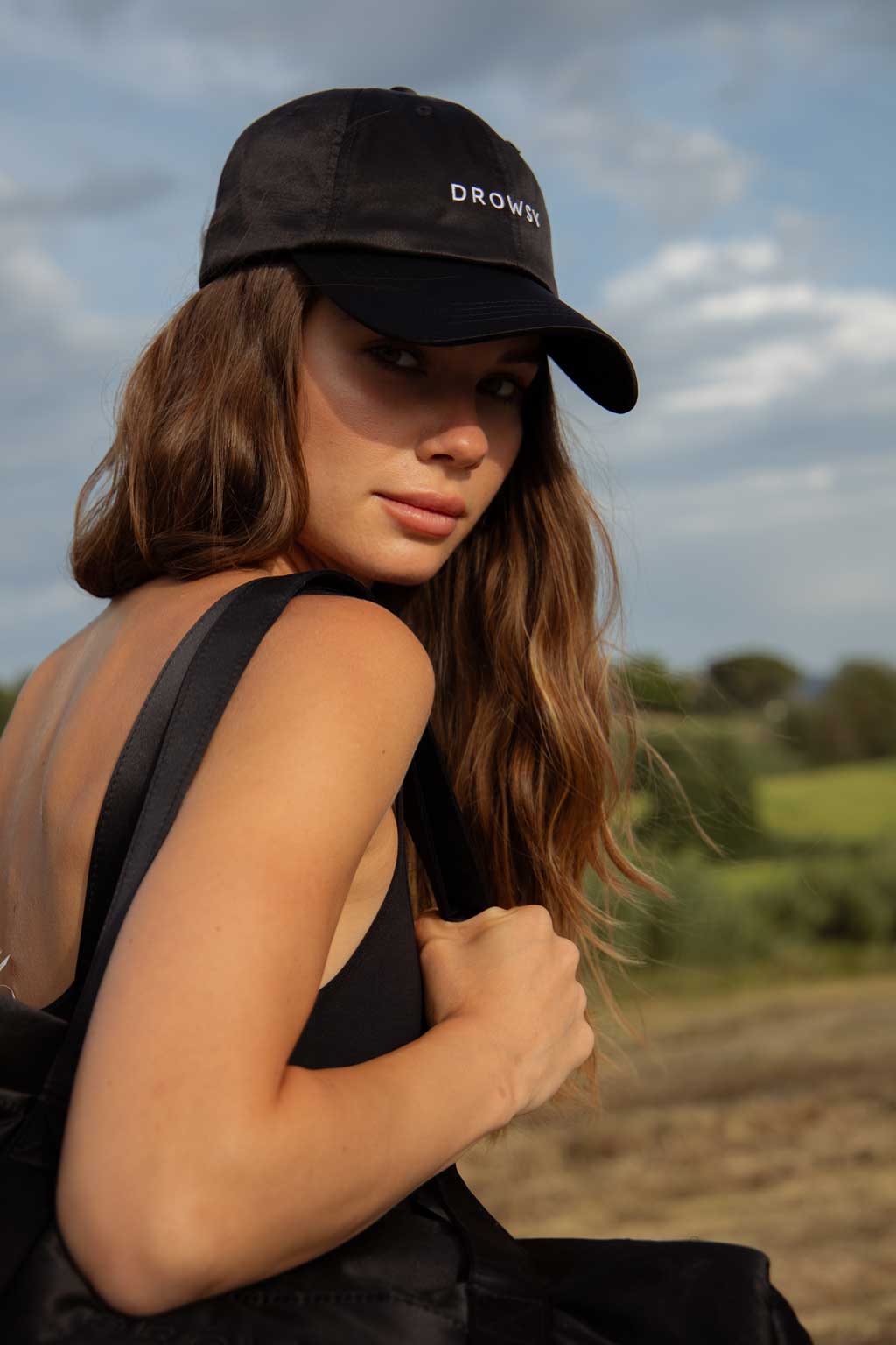 A woman with long brown hair, wearing a black cap and black tank top, looks over her shoulder outdoors in a natural setting with greenery and a cloudy sky in the background.