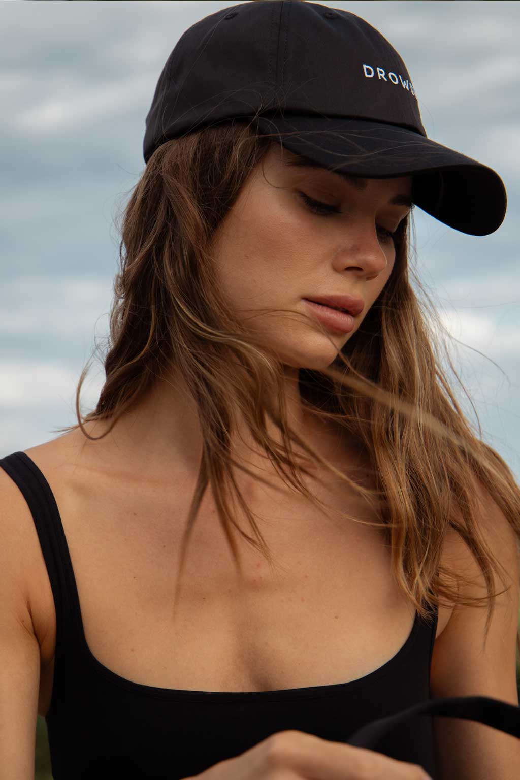 A young woman with long brown hair wears a black cap and black tank top, looking down with a serene expression. The background shows a cloudy sky.
