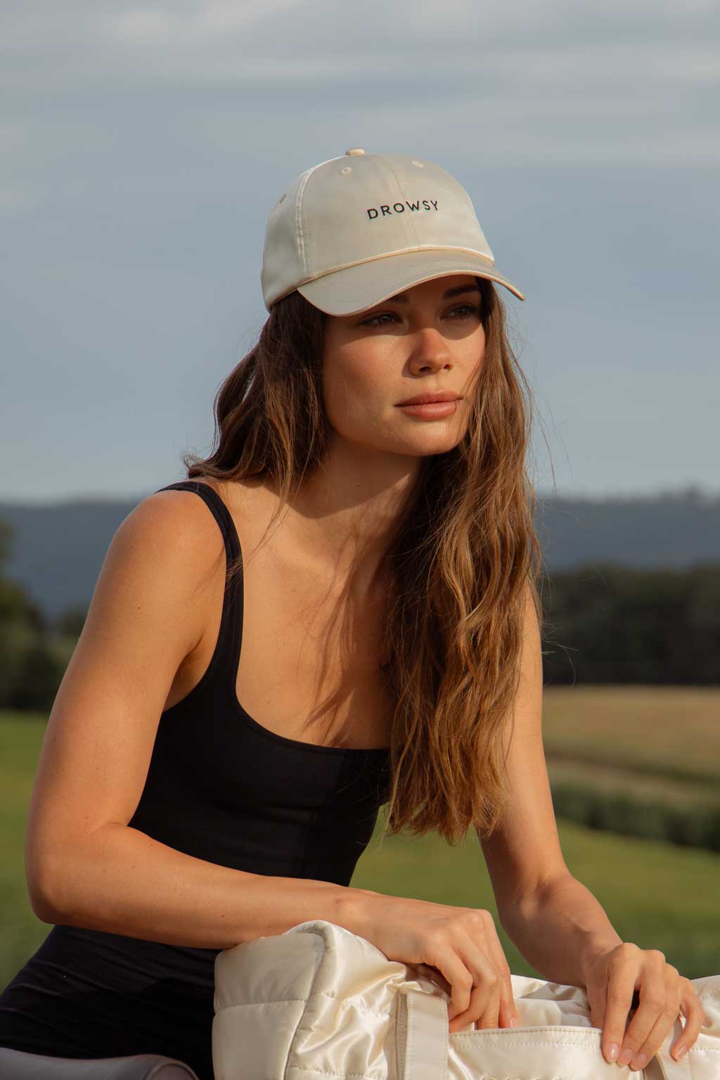 A woman with long brown hair wearing a beige cap and black tank top sits outdoors in a grassy field, holding a white bag. The background features rolling hills and a cloudy sky.