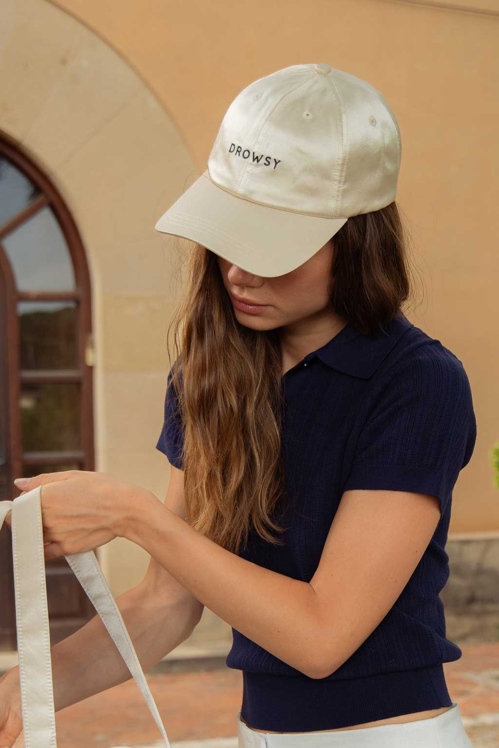 A woman with long brown hair wearing a beige baseball cap that says Drowsy, a navy short-sleeve shirt, and white pants holds a cream-colored strap outside near a building with an arched doorway.