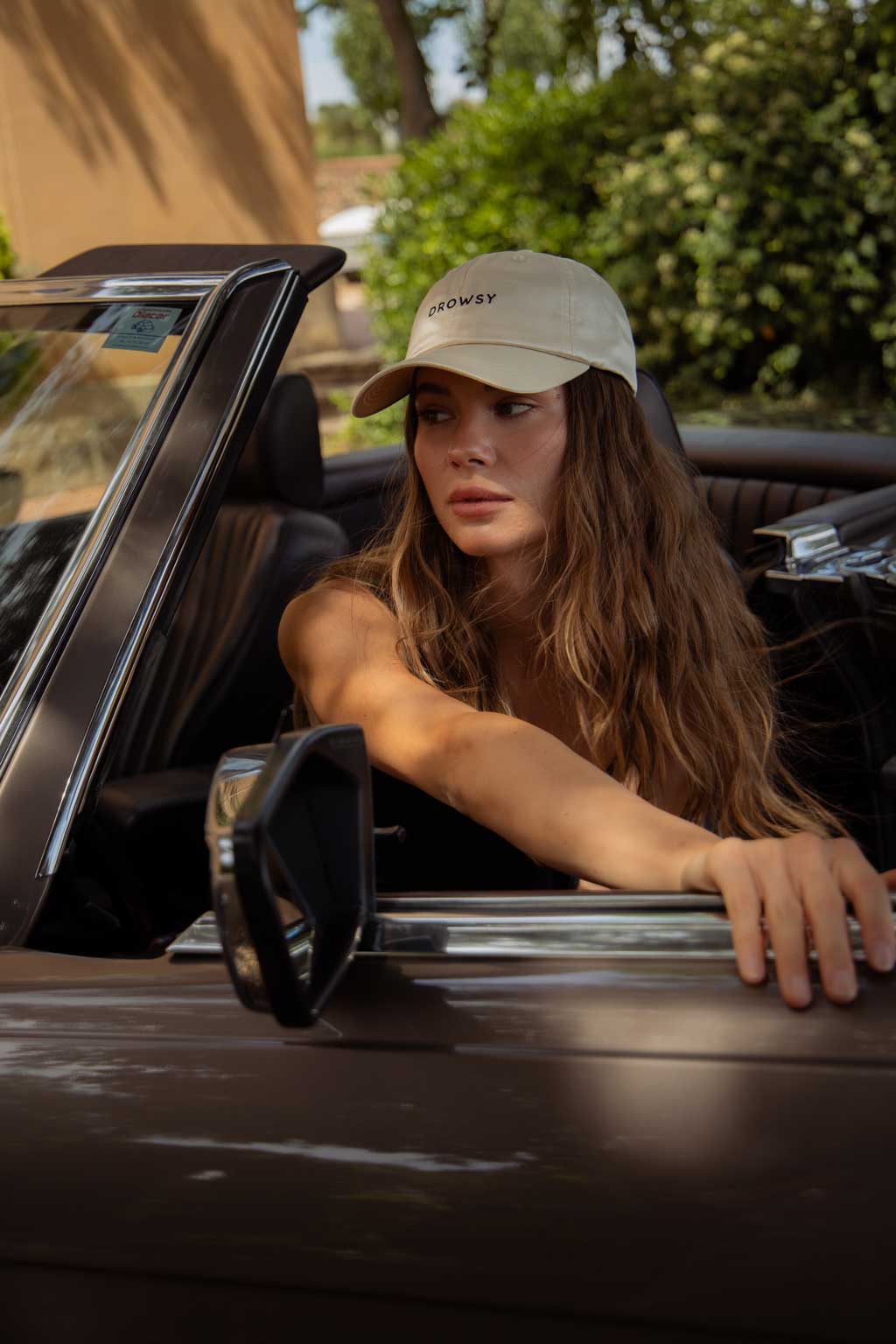 A woman with long wavy hair wearing a beige cap and black top sits in the driver’s seat of a convertible car, looking to her left with one hand on the steering wheel and the other on the car door. Trees and greenery are in the background.