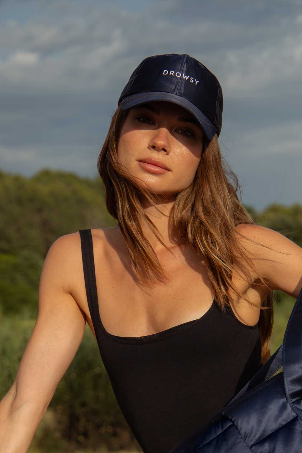 A woman with long brown hair wears a navy cap that says DROWSY and a black tank top, standing outdoors with trees and a cloudy sky in the background.