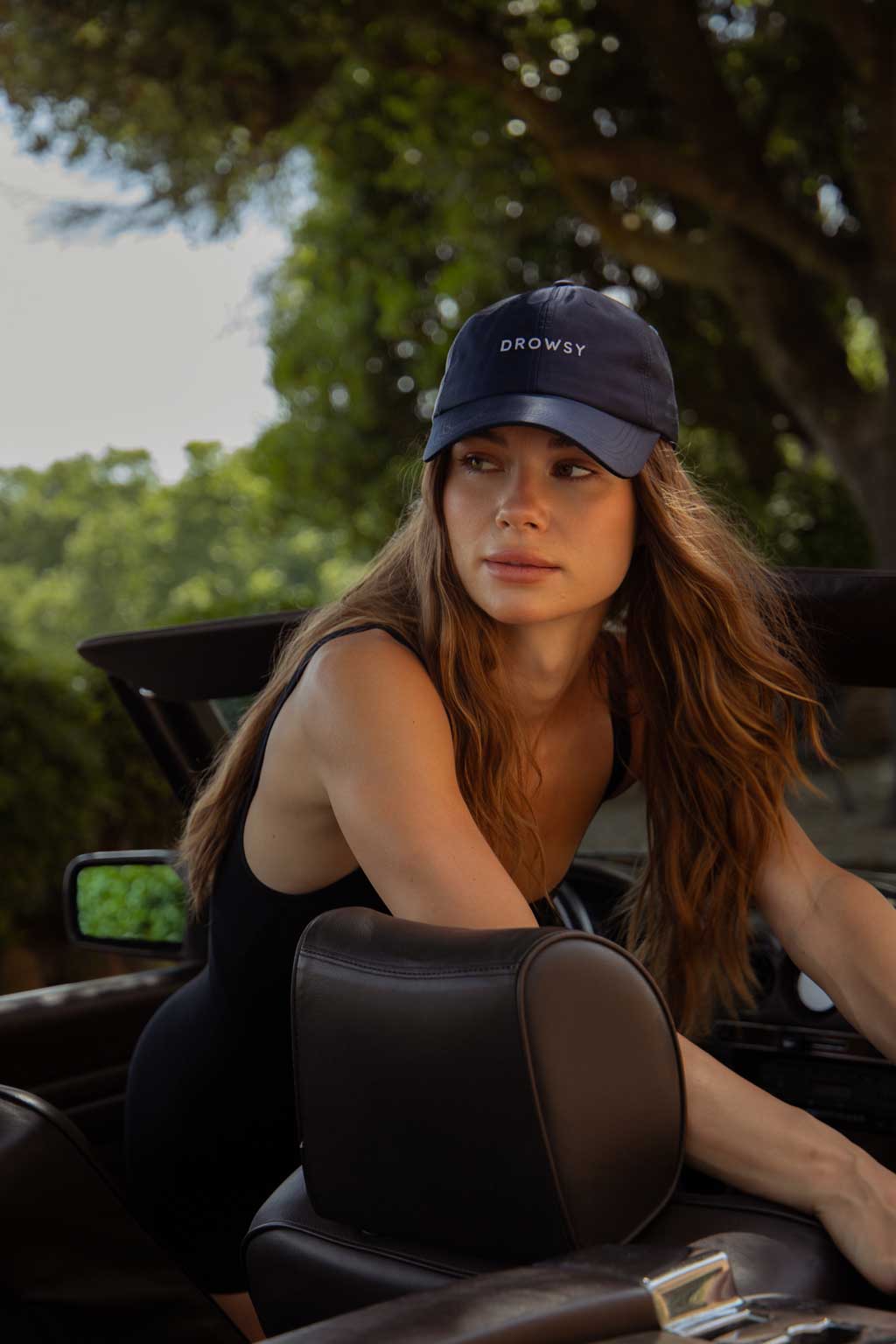 A woman with long brown hair, wearing a black sleeveless top and a navy cap that says DROWSY, sits in a convertible car with lush green trees in the background.