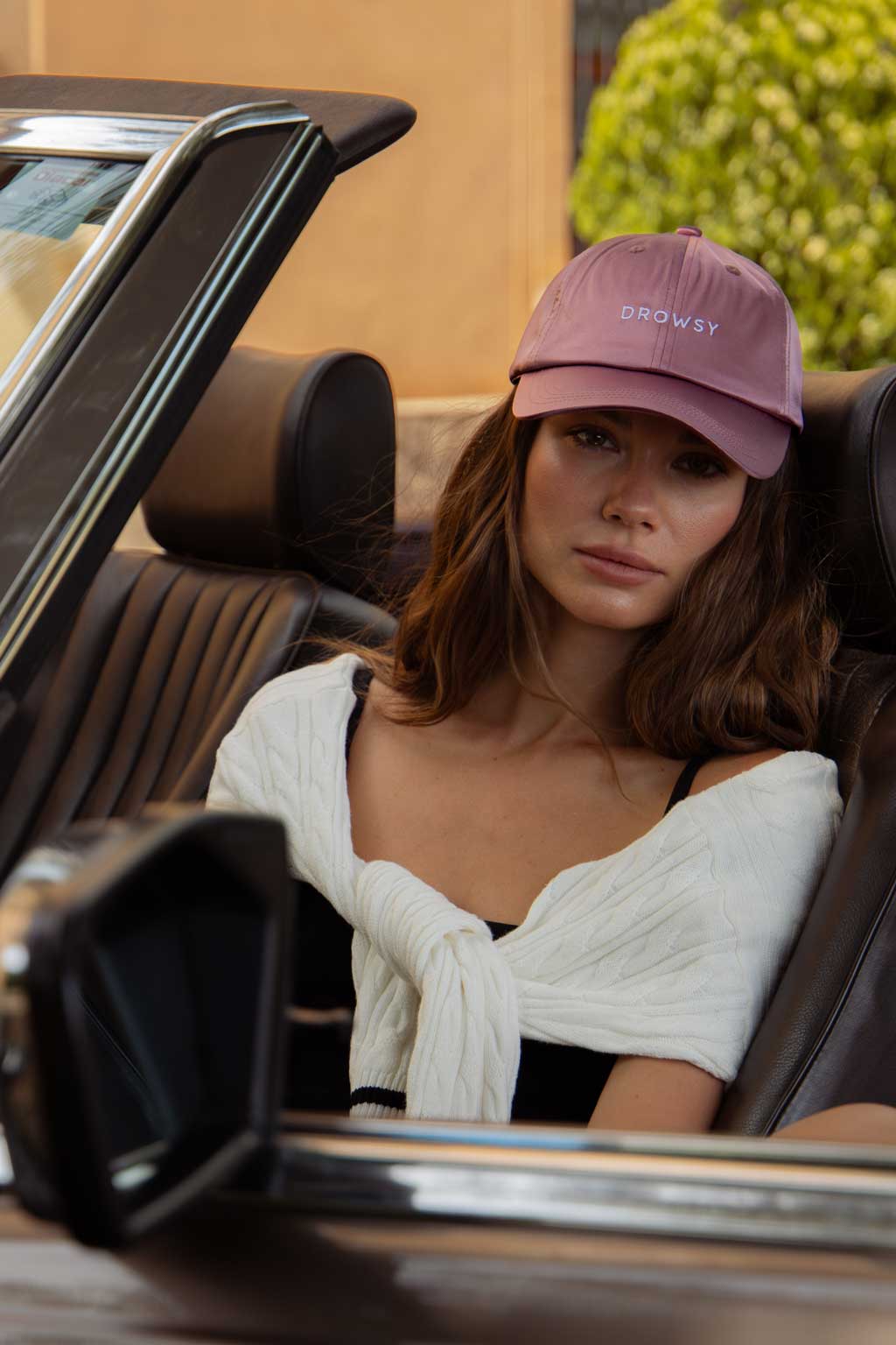 A woman wearing a pink cap that says “DROWSY” sits in the driver’s seat of a convertible car. She has a white sweater draped over her shoulders and looks calmly at the camera.