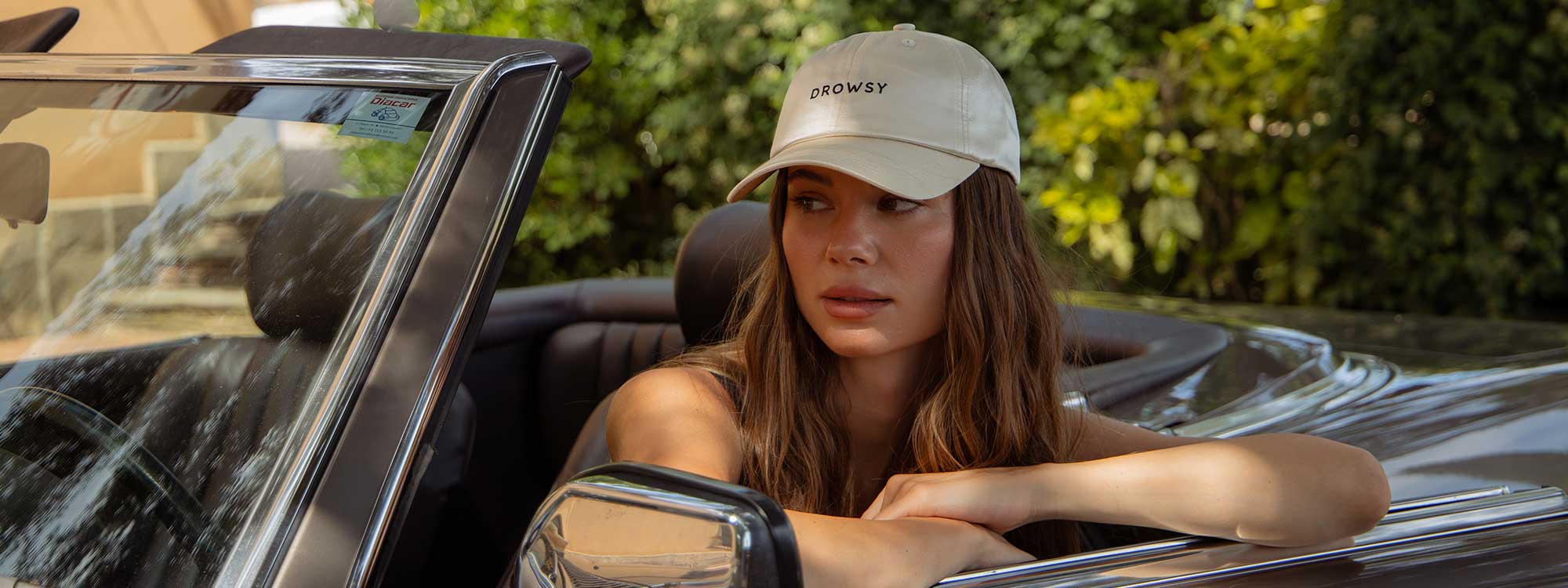 A woman with long brown hair wearing a beige DROWSY cap sits in a convertible car with the top down, resting her arms on the door, looking thoughtfully to the side. Green foliage is in the background.