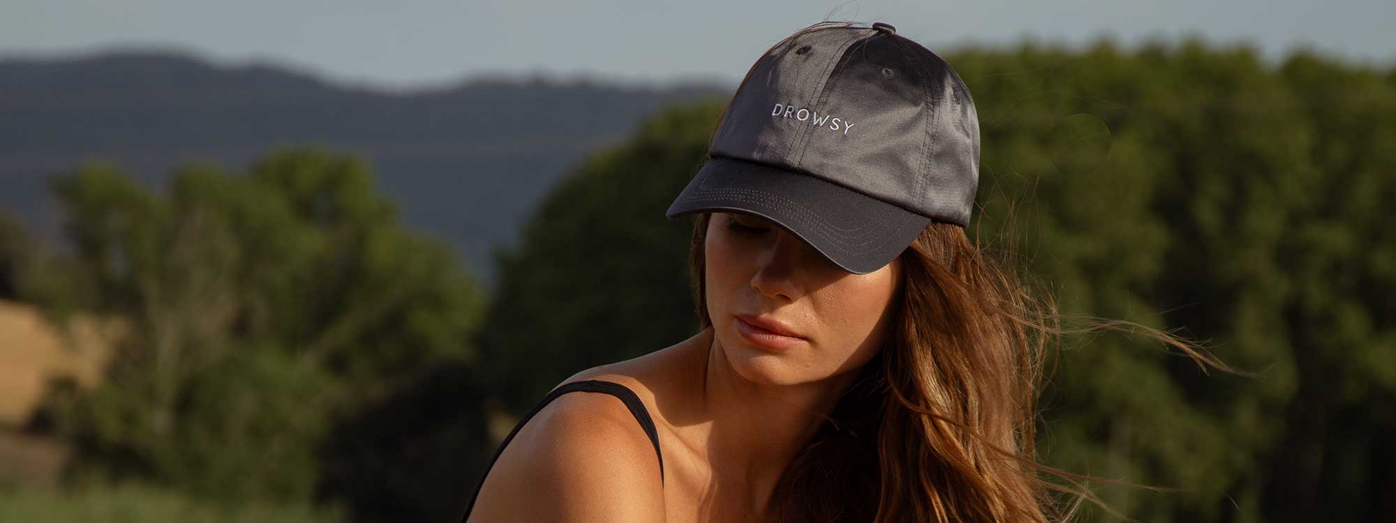 A woman wearing a black tank top and a gray baseball cap with the word DROWSY embroidered on it sits outdoors with trees and hills in the background.