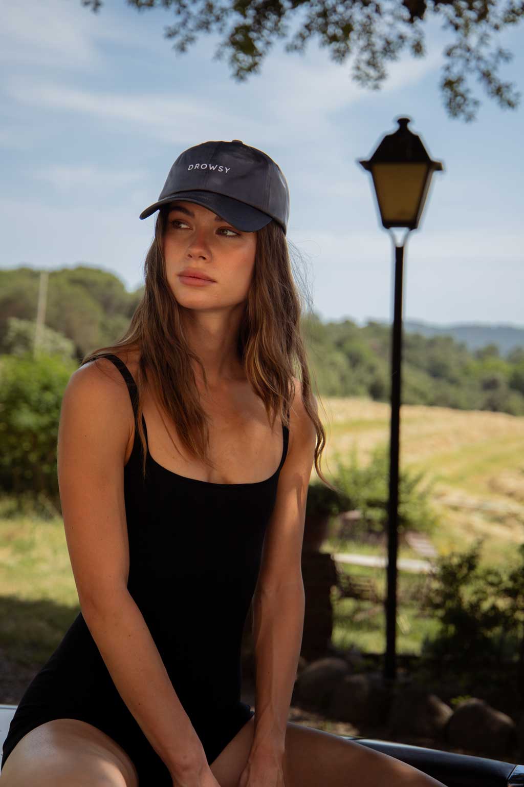 A woman with long brown hair in a black tank top and a dark cap sits outdoors, looking to the side. There is greenery, a lamp post, and a field in the background under a partly cloudy sky.