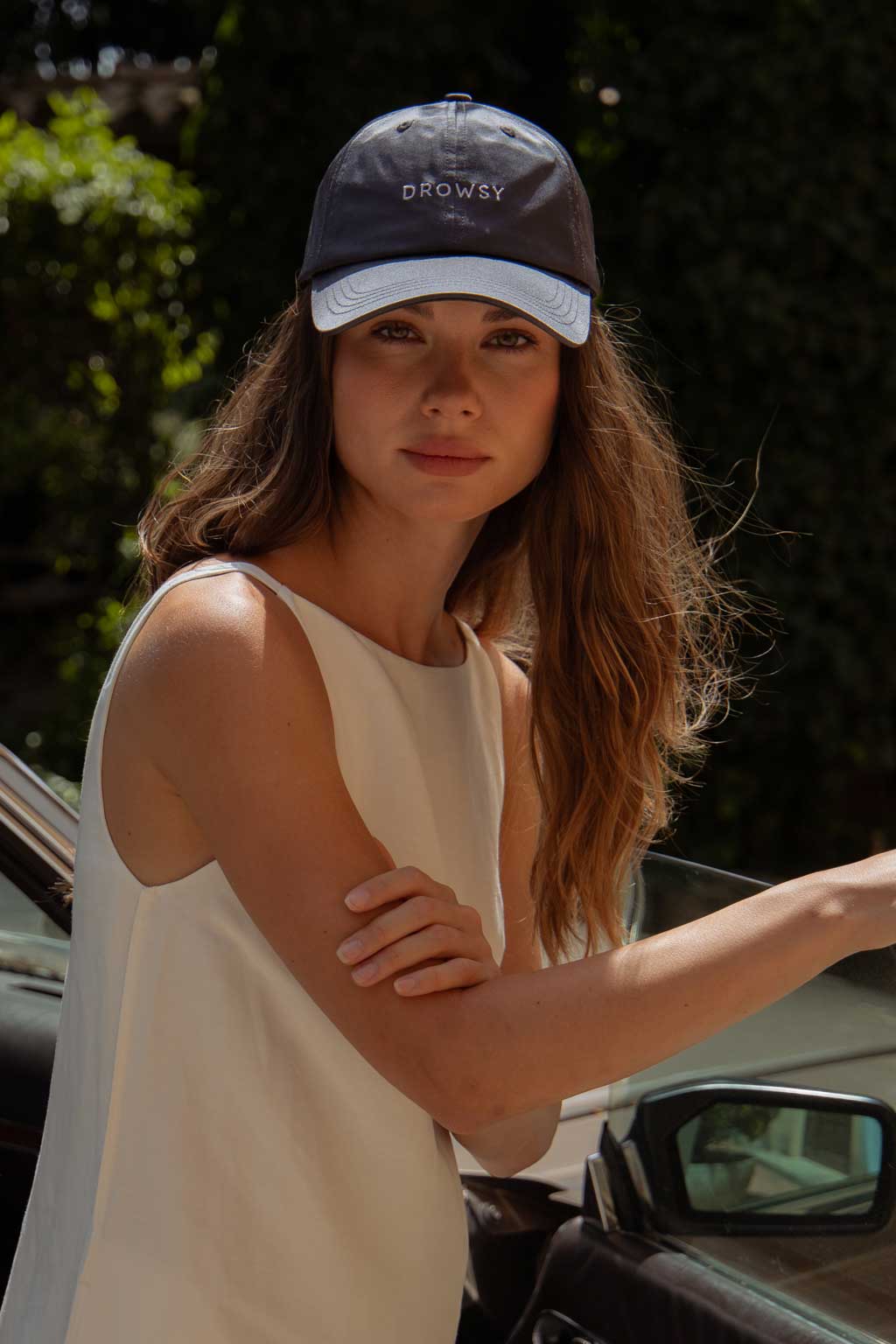 A woman with long wavy hair, wearing a sleeveless white top and a black cap that reads DROWSY, stands outdoors by a car, looking at the camera with a calm expression.